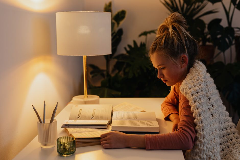 Young woman wrapped in a blanket reading under warm lamp light in a cozy room.