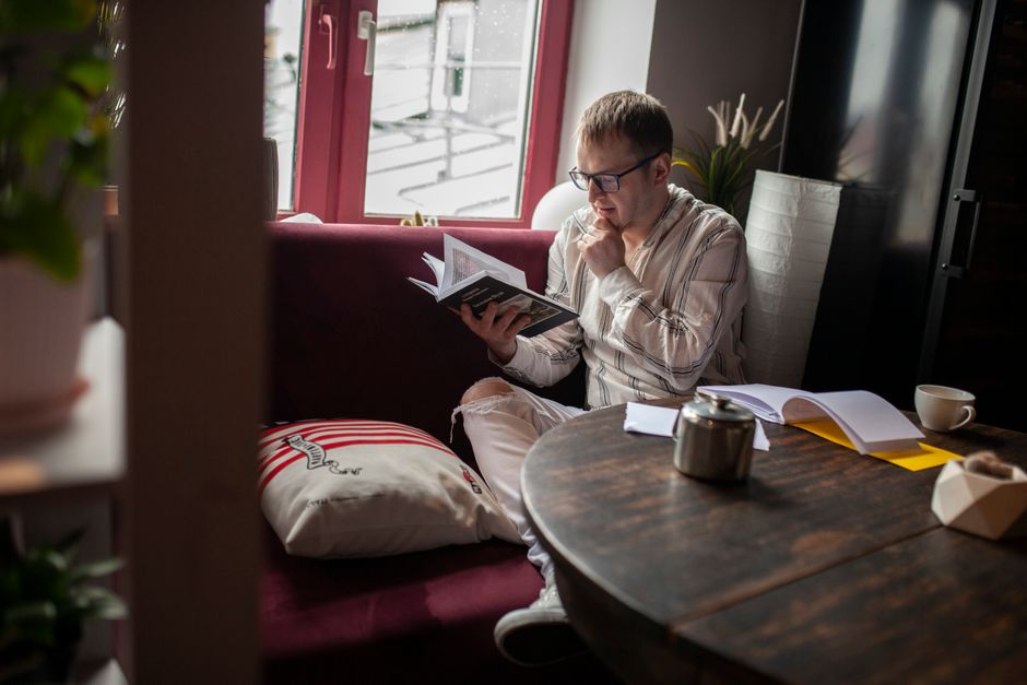 A man sits in a cozy room, reading a book by the window with a thoughtful expression.
