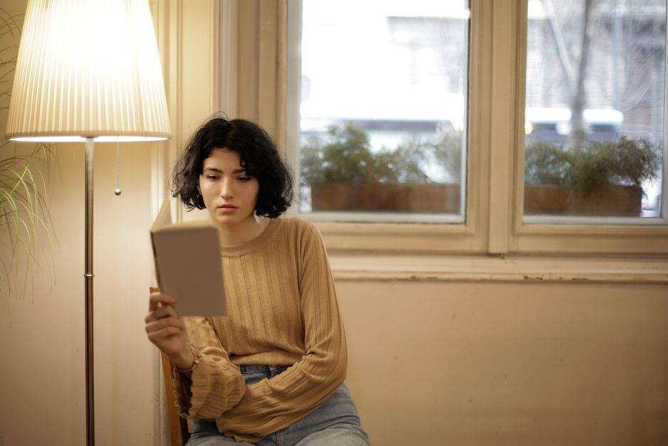 Woman sitting indoors reading near a window under warm lamplight.